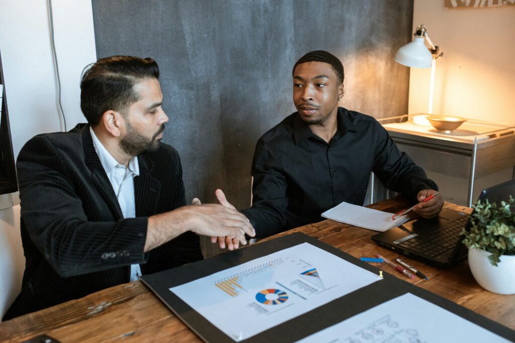 Two businessmen shaking hands over a desk with charts in a modern office.
